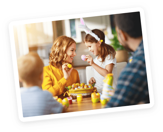 A woman and a girl wearing bunny ears smile and decorate Easter eggs at a table, while a man and a young child watch in a bright, cheerful room.