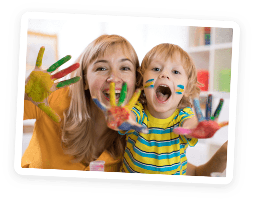 A smiling woman and a young child with painted hands and faces hold their hands toward the camera, enjoying a fun and colorful art activity indoors.