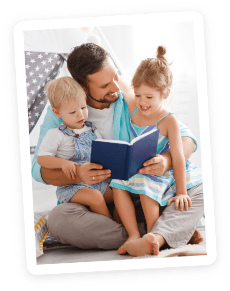 A bearded man sits on the floor reading a blue book to a young boy and girl. All three are smiling and appear happy, with a play tent visible in the background.