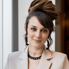 A woman with light skin, blue eyes, and brown hair styled in dreadlocks updo, wearing a beige blazer, black necklace, and earrings, smiles softly while standing indoors near a window.