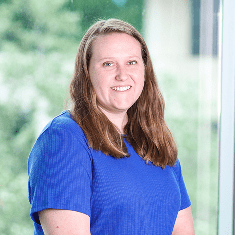 A woman with long brown hair wearing a blue shirt sits and smiles in front of a window with a blurred, green outdoor background.