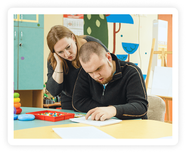 A woman leans over to assist a man who is focused on a workbook at a table in a colorful classroom, with educational toys and art supplies visible nearby.