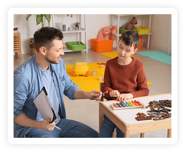 A man and a boy sit at a small table in a playroom. The man holds a clipboard and pen, while the boy plays with a colorful xylophone. Wooden puzzle pieces are on the table, and toys are scattered in the background.