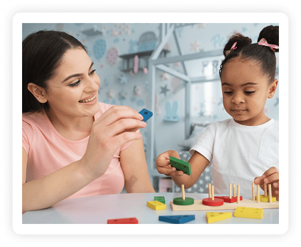 A woman and a young girl sit at a table, smiling and playing with colorful wooden shape sorting toys in a bright, decorated room.