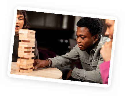 Three people sit around a table, focused on playing a game of Jenga. One person carefully pulls a wooden block from the tower while the others watch attentively.