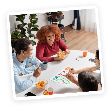 Four people sit around a white table playing a colorful board game, smiling and talking. Snacks and drinks are on the table, and a potted plant is in the background.