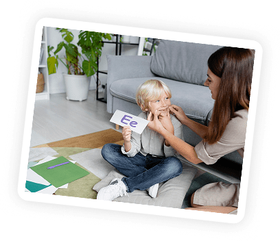 A woman teaches a young boy the letter "E" using a flashcard while sitting on the floor in a cozy living room with books and folders nearby.