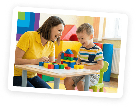 A woman and a young boy sit at a small table, smiling and playing with colorful building blocks in a bright, cheerful room.