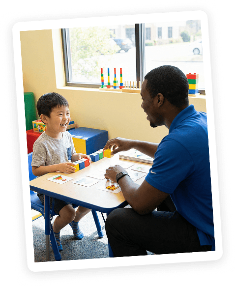 A young boy and a teacher sit at a small table, smiling and playing with colorful building blocks in a bright classroom with sunlight coming through the window.
