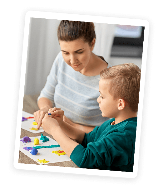A woman and a young boy sit at a table making colorful shapes with modeling clay, smiling and working together on a creative project.