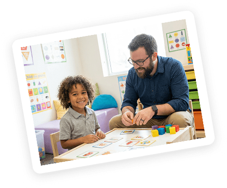 A young child and an adult sit at a classroom table, smiling and playing with colorful blocks and cards. The room has educational posters and a bright, welcoming atmosphere.