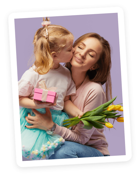 A young girl kisses a smiling woman on the cheek. The girl holds a small pink gift box and wears a crown, while the woman holds a bouquet of yellow tulips. Both are against a purple background.