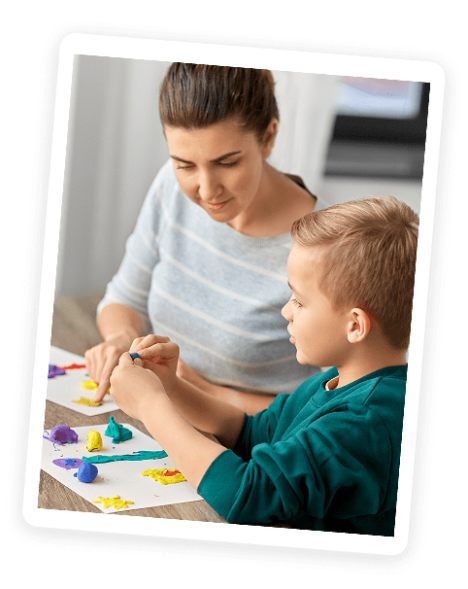 A woman and a young boy sit at a table making colorful shapes with modeling clay, smiling and working together on a creative project.